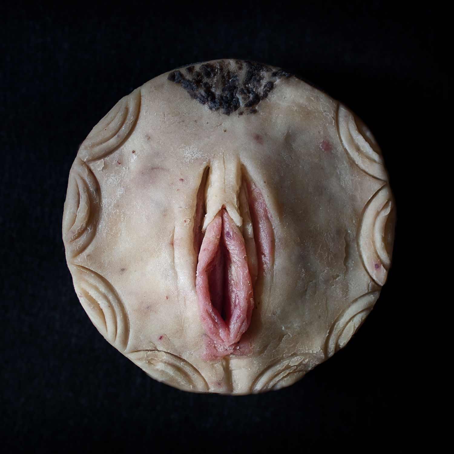 Real pie crust with pie art that looks like a vulva with pink inner labia and a small amount of pubic hair above the clitoral hood.
