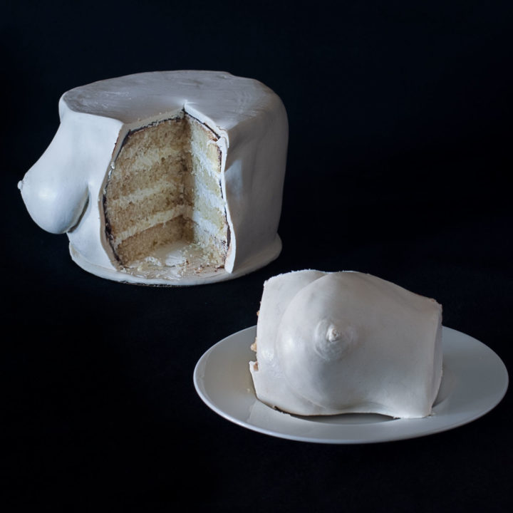Hand sculpted buttermilk cake with Swiss meringue buttercream. Cake is sculpted to look like realistic breasts. One breast is sliced out of the cake and on a plate on the lower right side of the image. The sliced cake in the background.