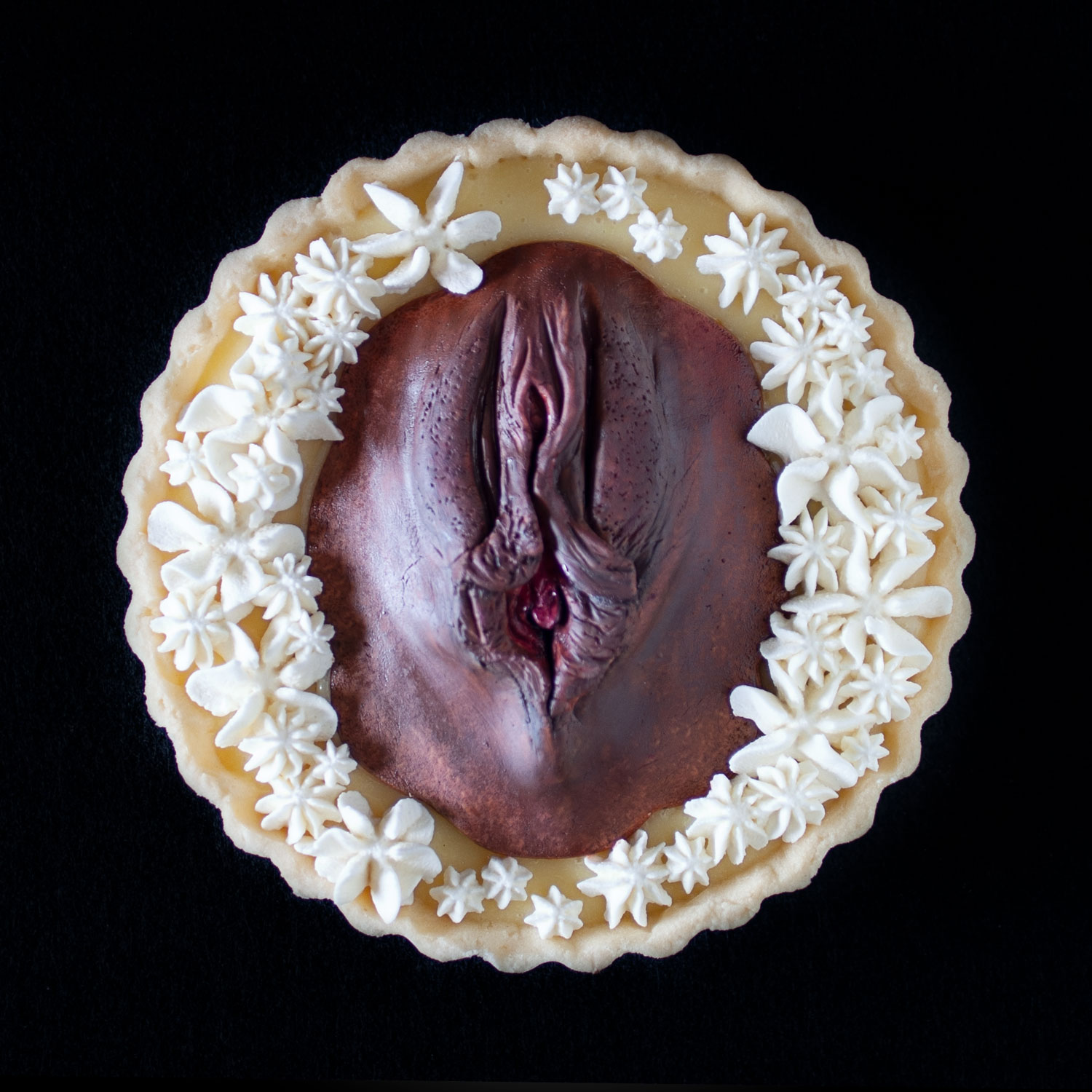 A top view of a lemon cream tart decorated with piped whipped cream and a hand sculpted vulva painted to look like a realistic human vulva with dark pigmented skin