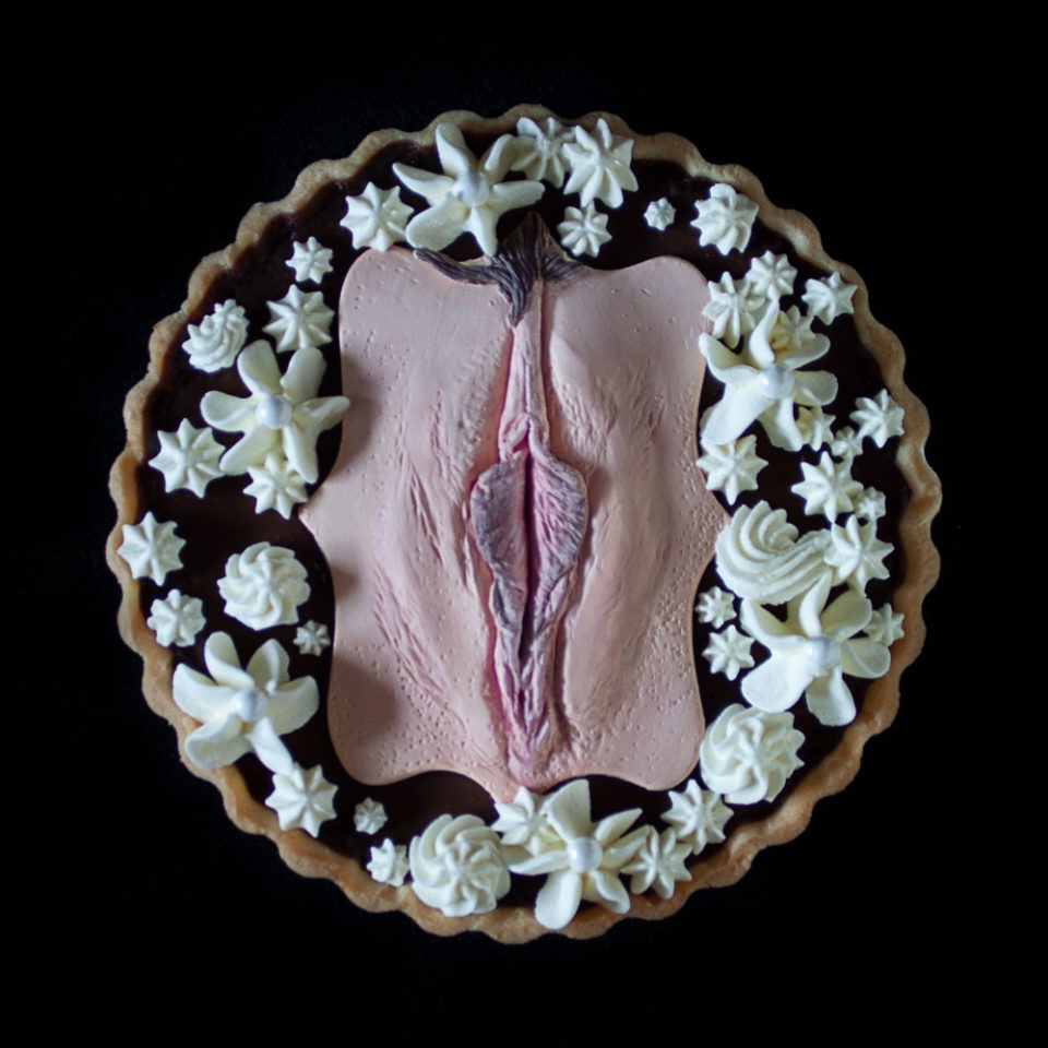 A French Chocolate Orange tart on a black background. A hand sculpted vulva art made of chocolate sits in the center of the tart with piped whipped cream surrounding the vulva.