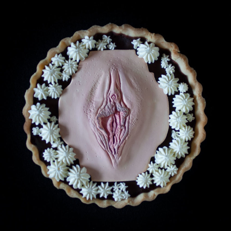 A French Chocolate Orange tart on a black background. A hand sculpted vulva art made of chocolate sits in the center of the tart with piped whipped cream surrounding the vulva.