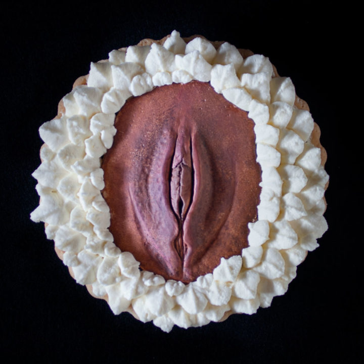 A French Chocolate Orange tart on a black background. A hand sculpted vulva art made of chocolate sits in the center of the tart with piped whipped cream surrounding the vulva.
