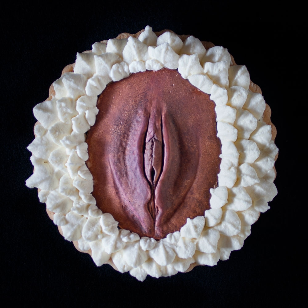 A French Chocolate Orange tart on a black background. A hand sculpted vulva art made of chocolate sits in the center of the tart with piped whipped cream surrounding the vulva.