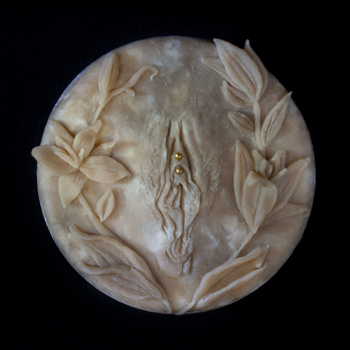 Hand sculpted pie crust art with pie crust vulva surrounded by a wreath of pie crust lilies on a black background. The clitoral hood appears pierced with gold dragees