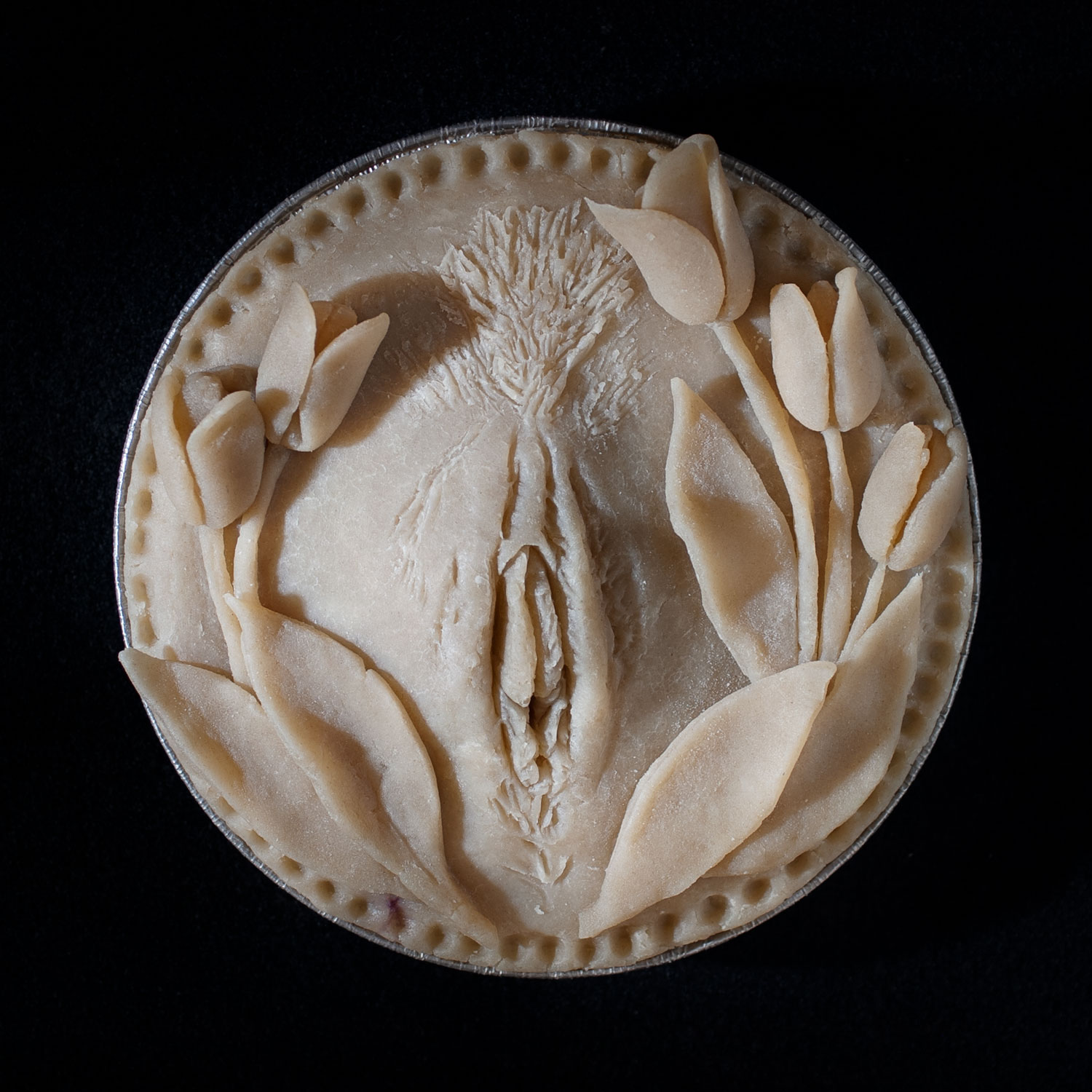 An unbaked pie on a black background. The hand sculpted pie crust art has a vulva in the center surrounded by a wreath of tulips.
