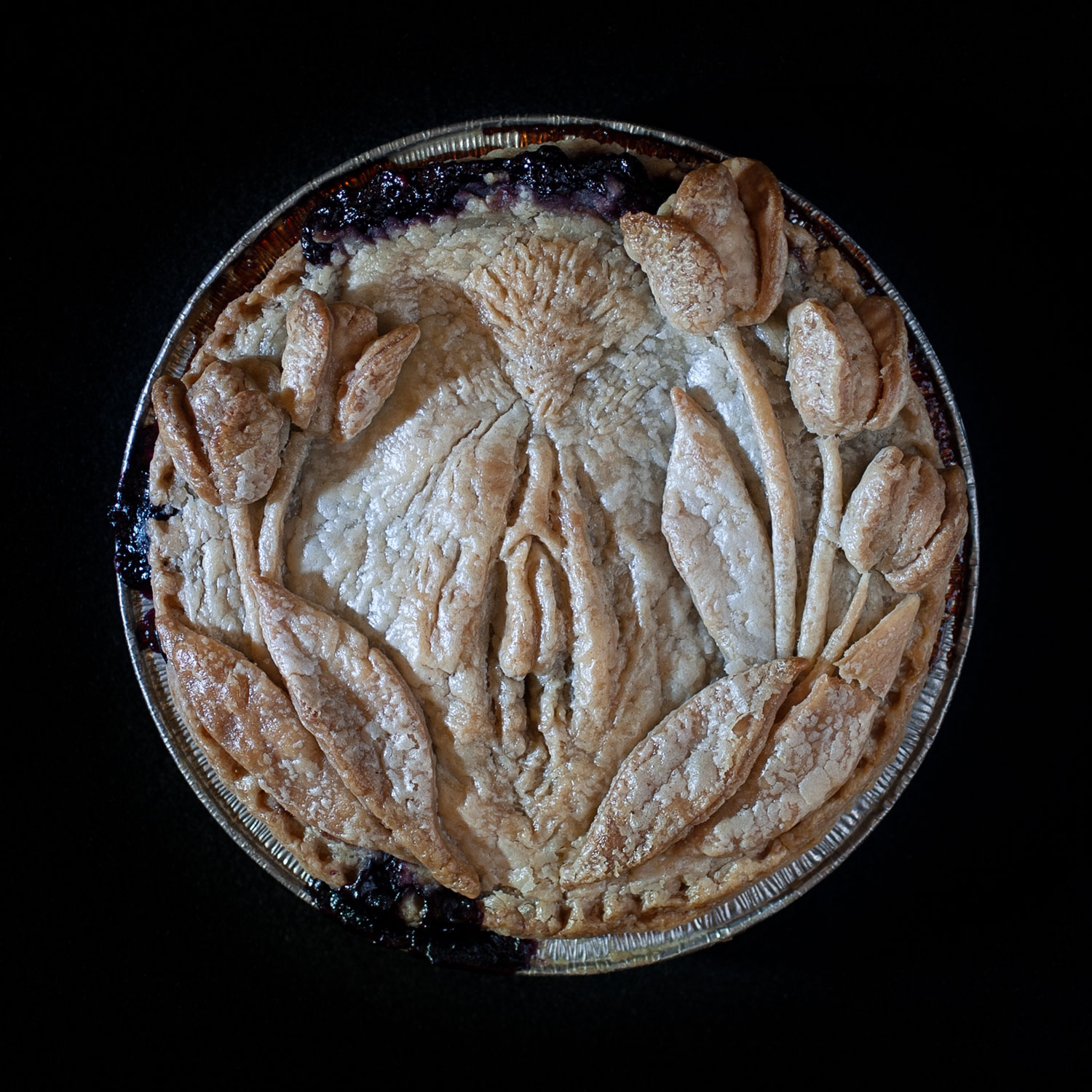 A baked pie on a black background. The hand sculpted pie crust art has a vulva in the center surrounded by a wreath of tulips. The blueberry filling has bubbled out of the top of the pie crust.