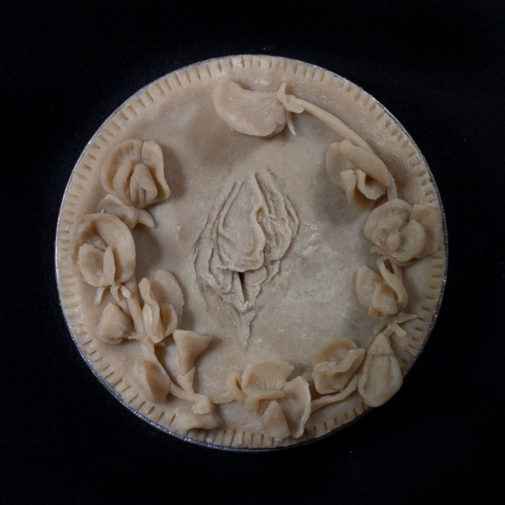 Unbaked pie with hand sculpted pie crust art. The pie features vulva art in the center surrounded by sculpted sweet pea flowers.