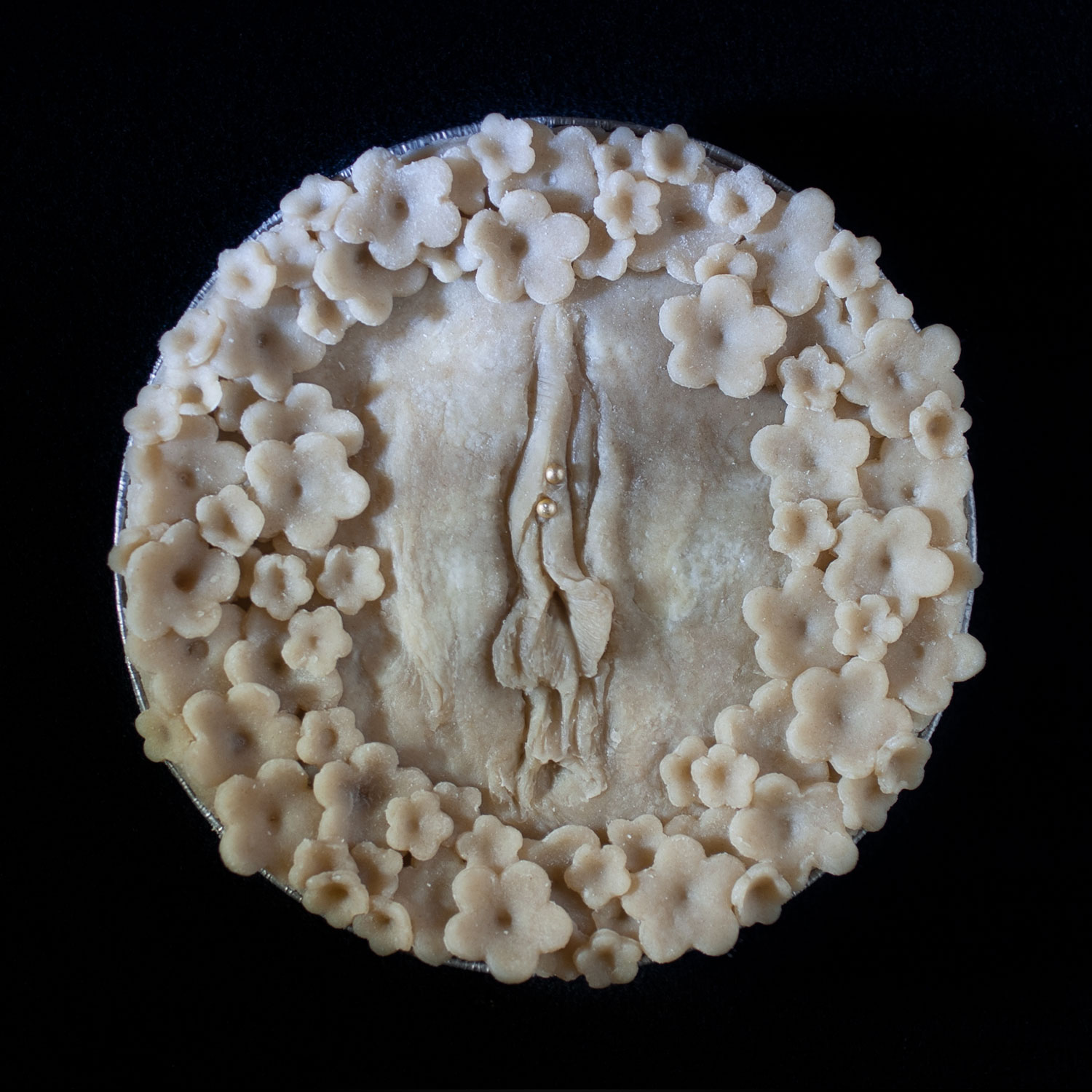 An unbaked artistic pie on a black background. The pie is decorated with hand sculpted vulva art and blossoms surrounding it. 