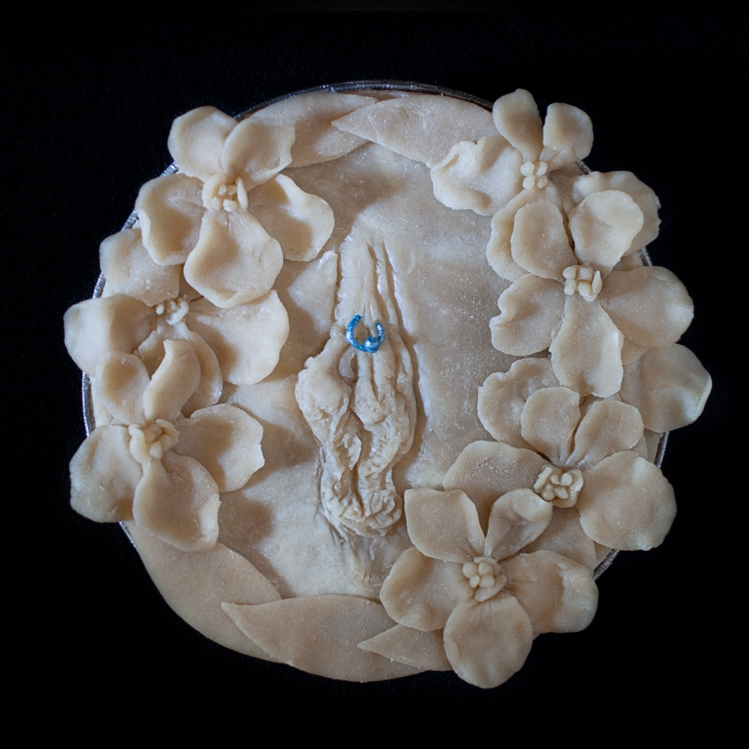 Unbaked pie on a black background. The pie crust art features a wreath of hand made violets and a pierced vulva with a blue clitoral hood piercing.