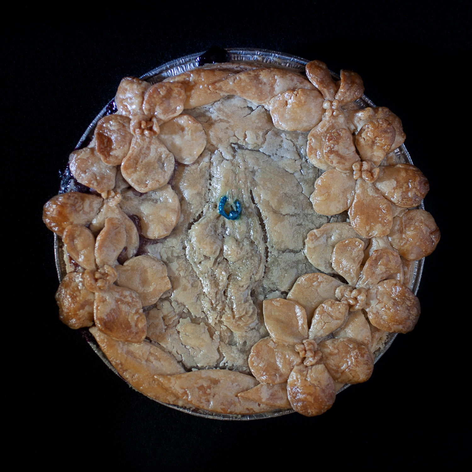Baked pie on a black background. The pie crust art features a wreath of hand made violets and a pierced vulva with a blue clitoral hood piercing.