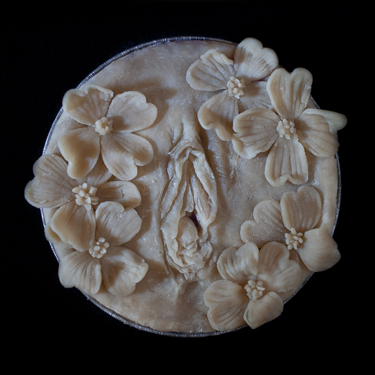 Unbaked pie on a black background. In the center of the pie is a hand sculpted pie crust vulva surrounded by dogwood flowers made of pie dough.