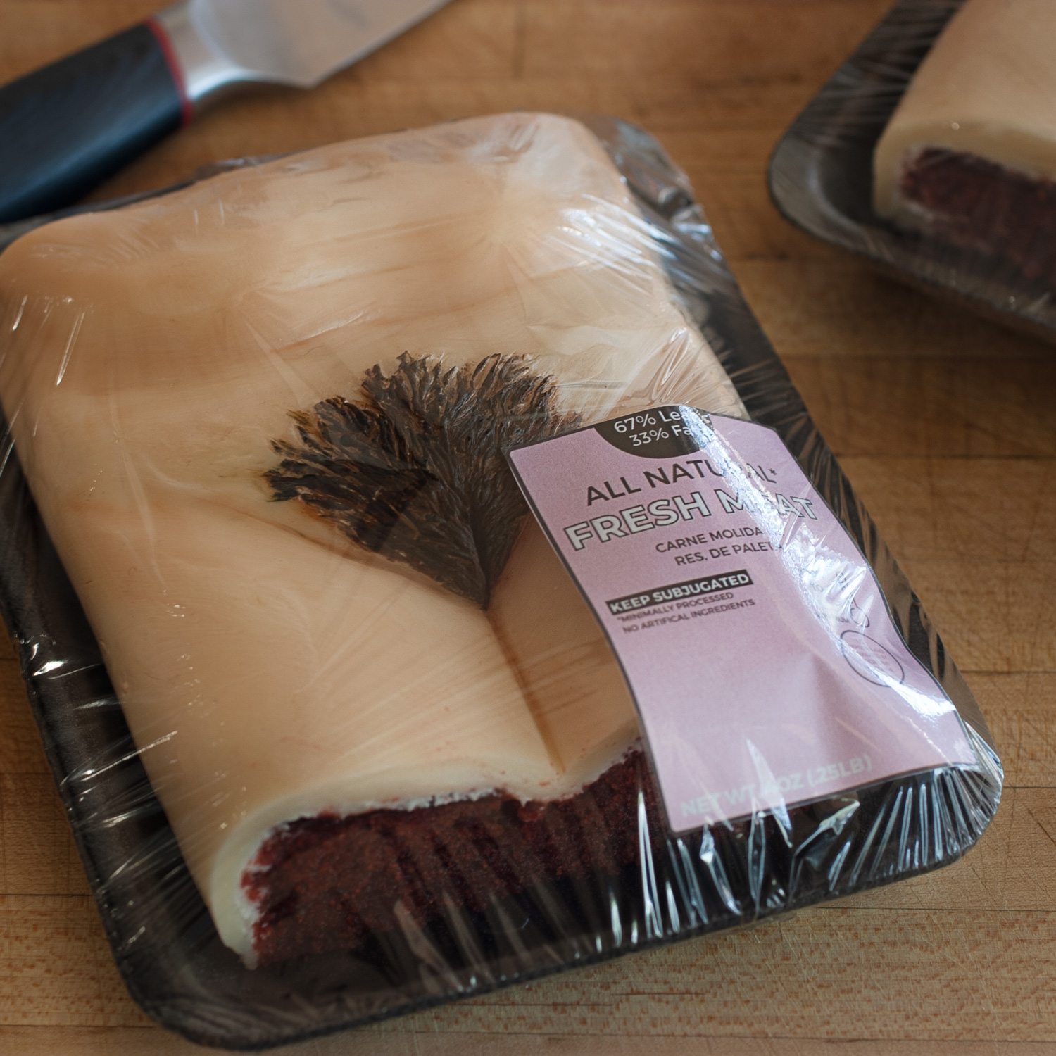 Red velvet cake with modeling chocolate art made to look like a human vulva with a brown triangle of pubic hair. The cake is packaged in a styrofoam meat tray, wrapped in plastic wrap with a pink label.