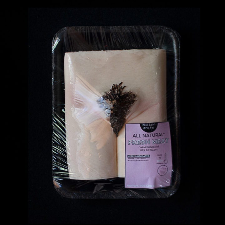 art portraying a human vulva wrapped in plastic and packaged like fresh meat with a pink label sitting on a black background