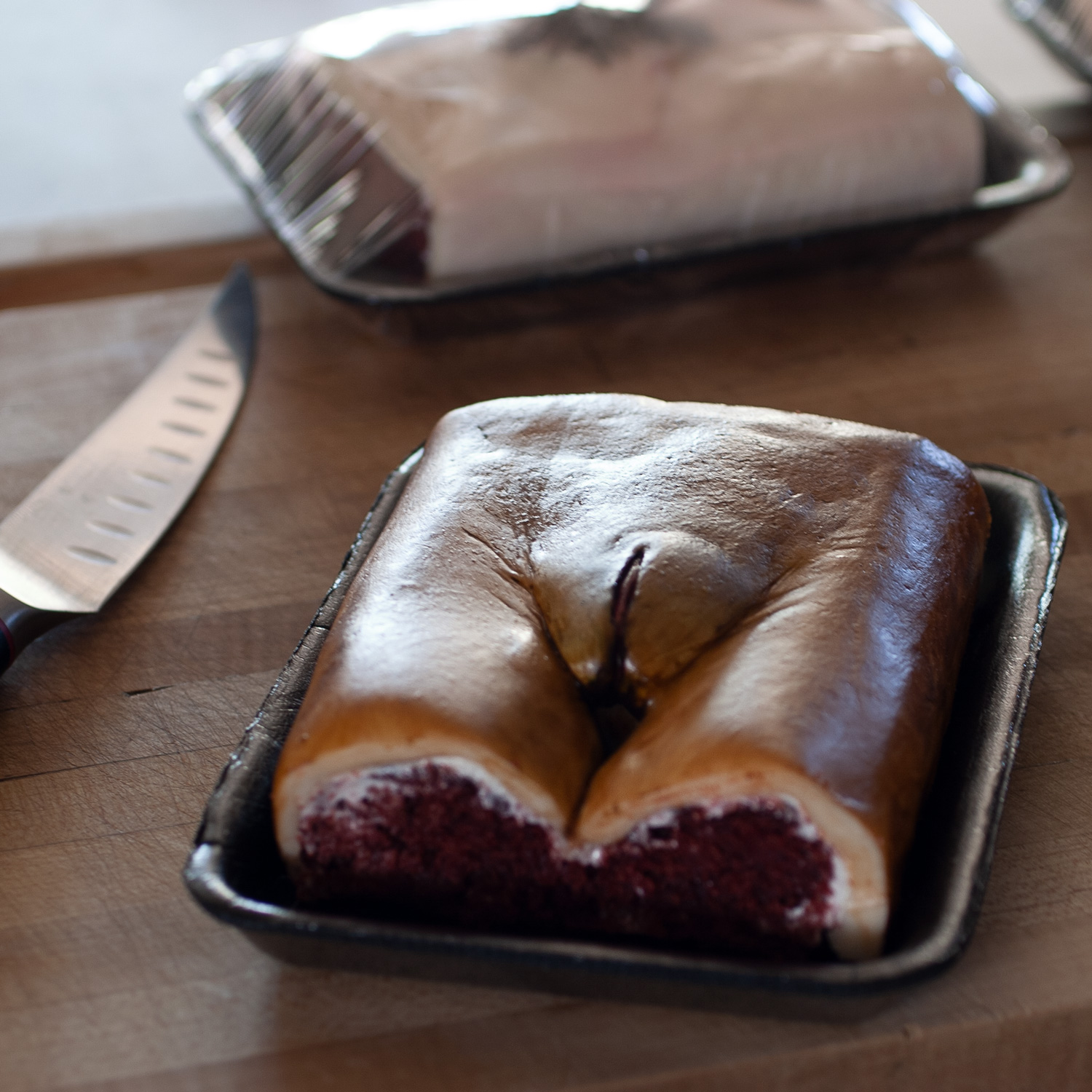 Red velvet cake in a black styrofoam meat tray. The cake is decorated to look like a realistic human vulva with brown skin. In the background is another vulva cake and a boning knife. 