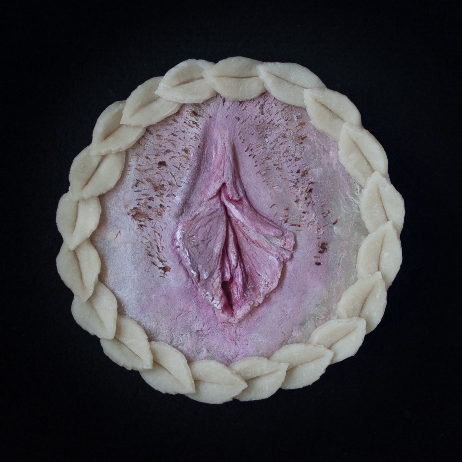 Mini vulva pie on black background with hand sculpted, hand painted vulva art surrounded with pie crust leaves