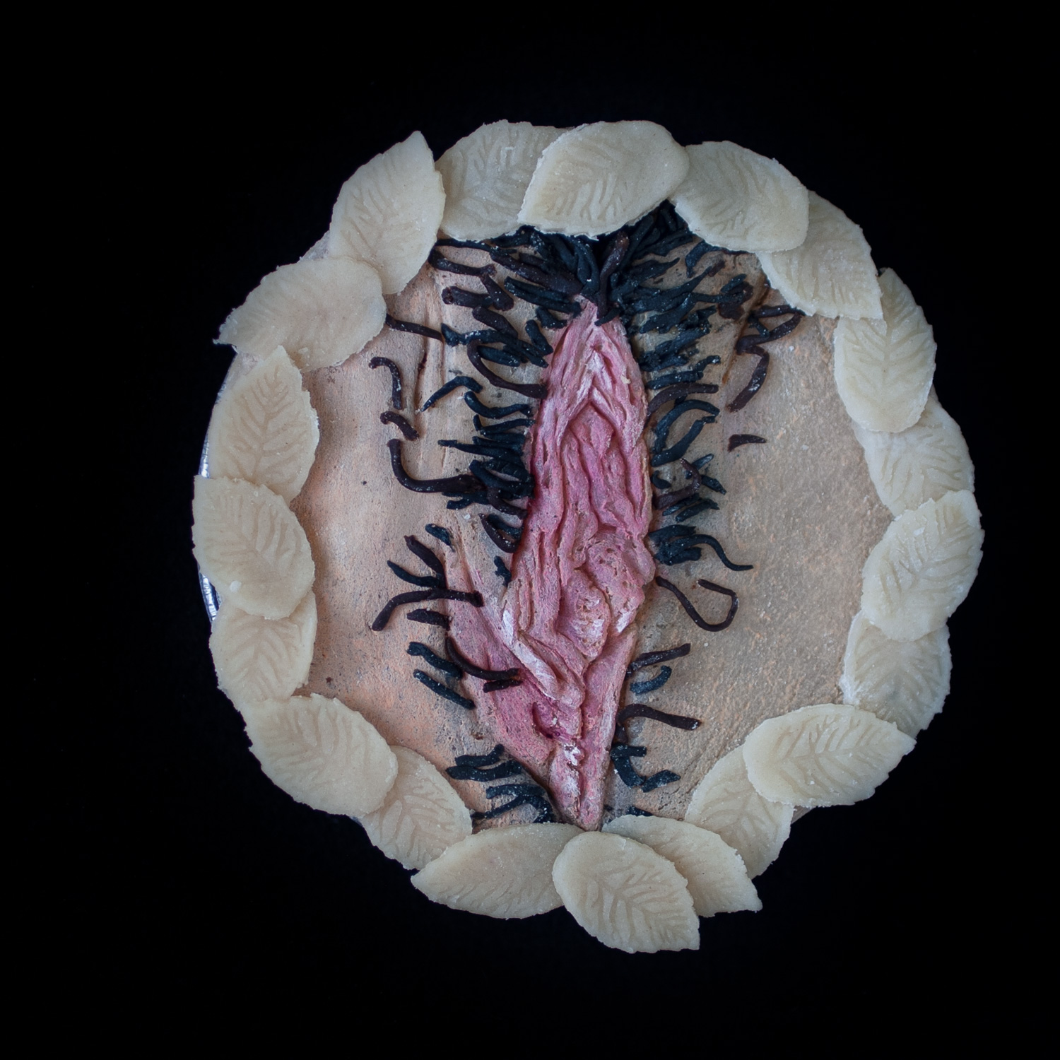 A vulva pie for Pies in the Window on a black background. The Pie features hand-sculpted pie crust art and pubic hair surrounded my pie dough leaves. 