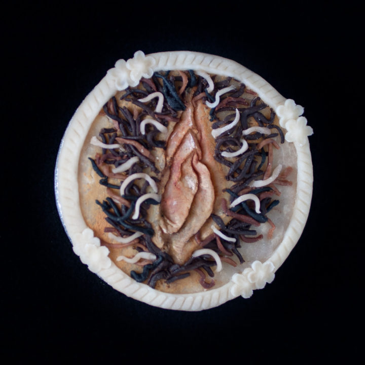 Pie on black background with realistic vulva art made from multicolored pie dough.