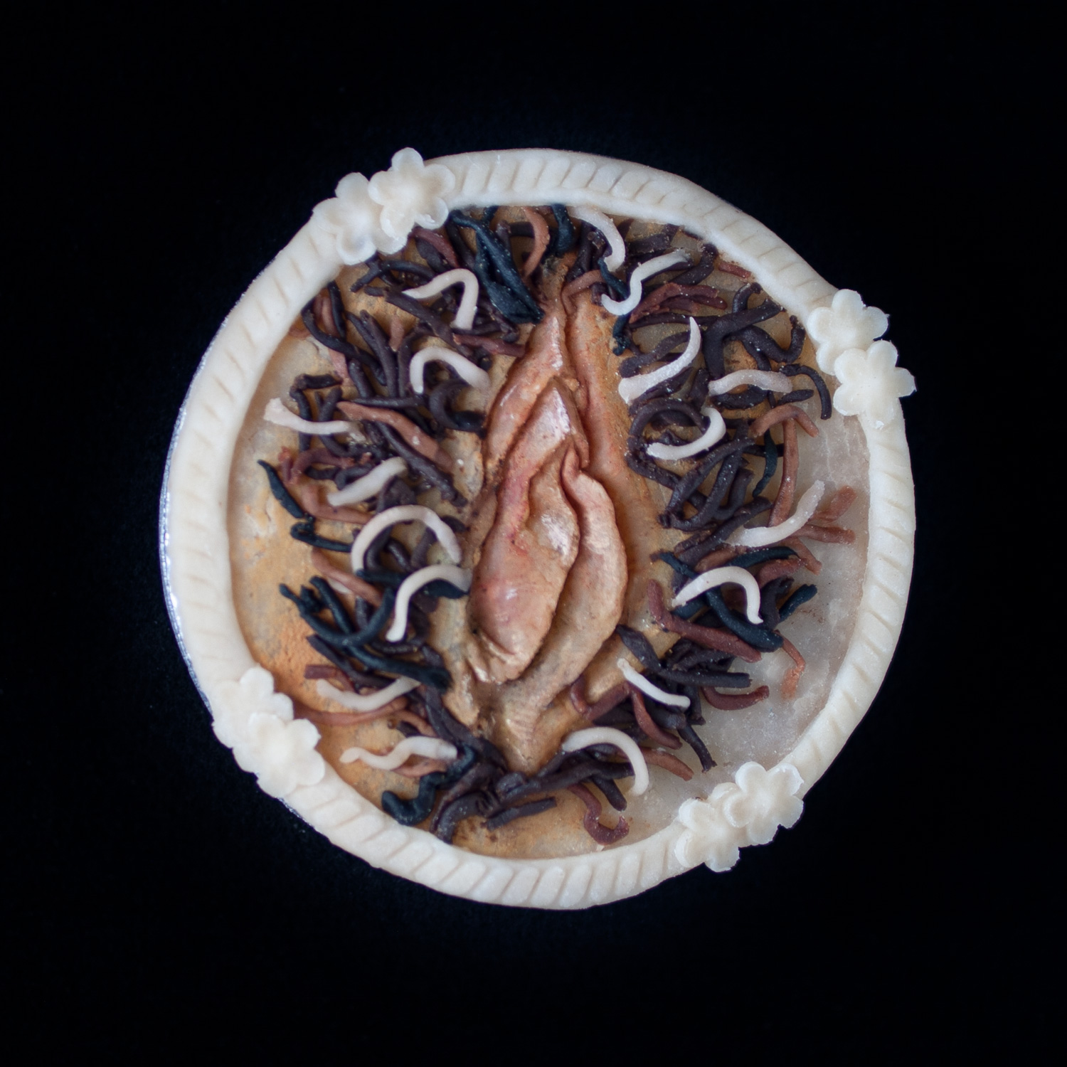 Pie on black background with realistic vulva art made from multicolored pie dough. 