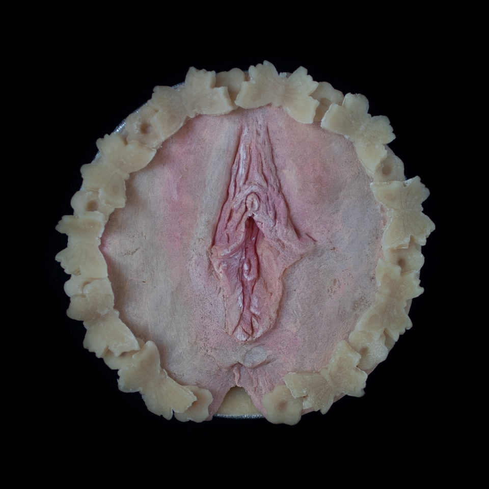 A pie on a black border with hand sculpted vulva made of pie dough.