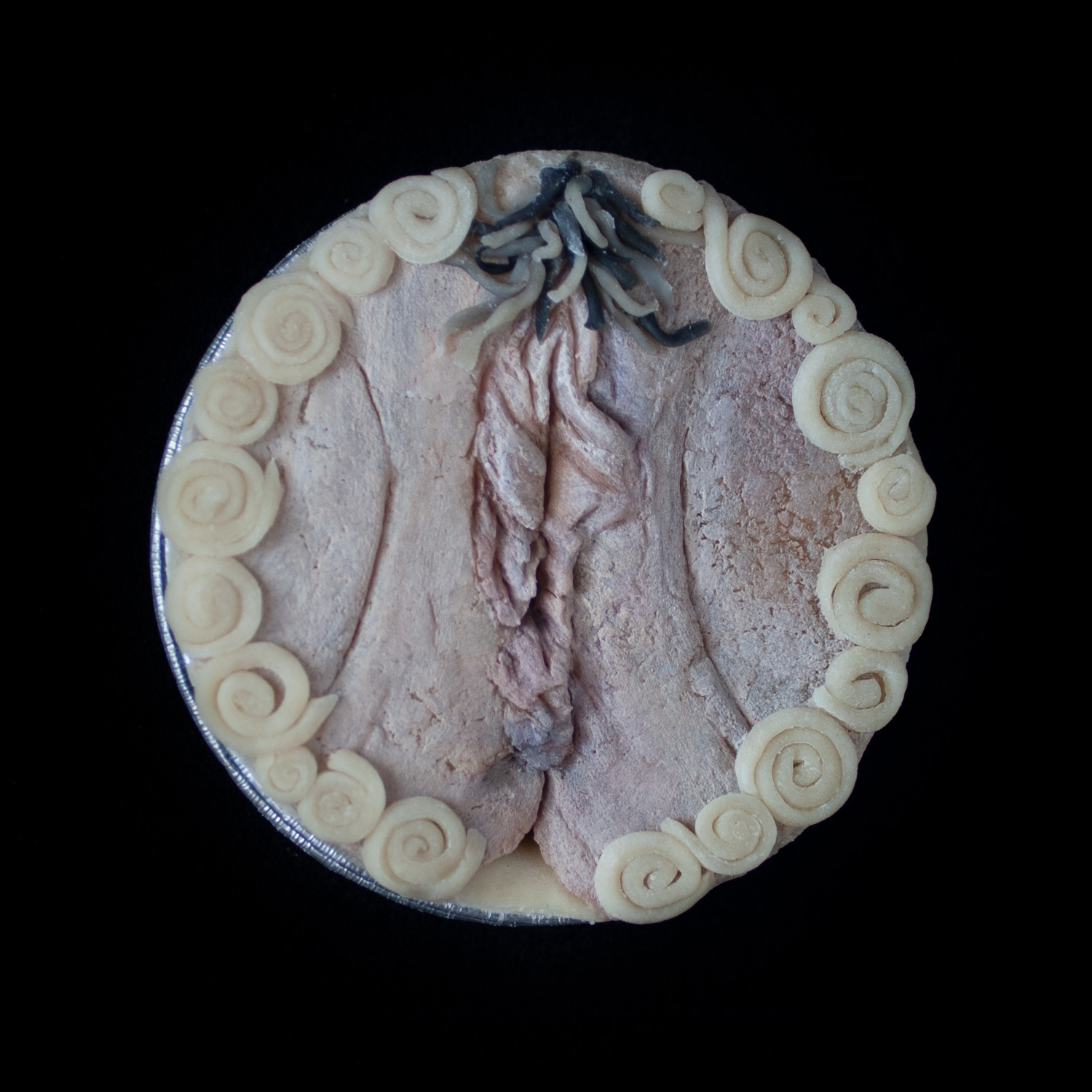 Mini-pie on a black background decorated with hand sculpted, hand painted vulva art surrounded with spiral border