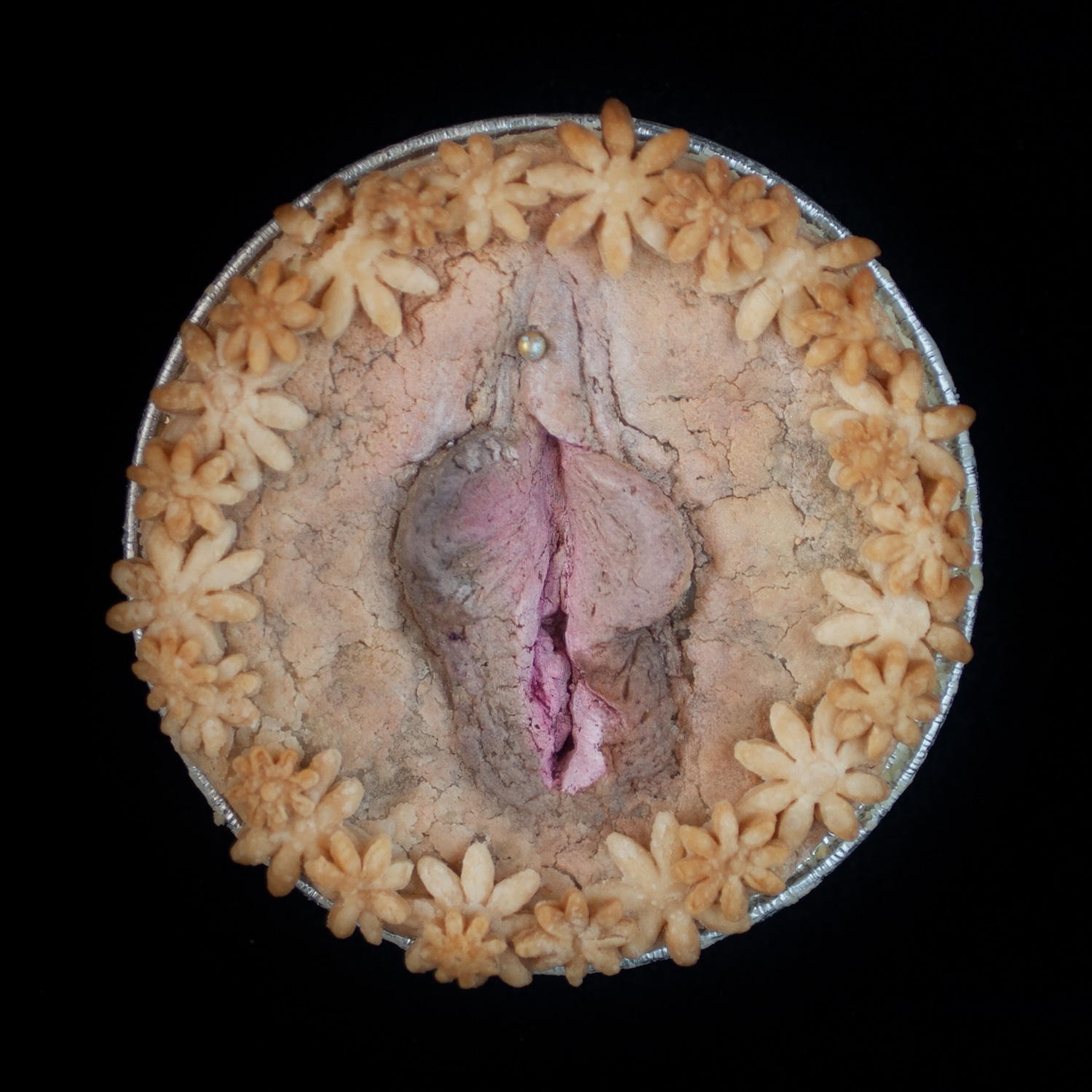 Baked pie on black background, the pie is decorated with pie crust flowers and a pie crust vulva made in a very realist fashion. 