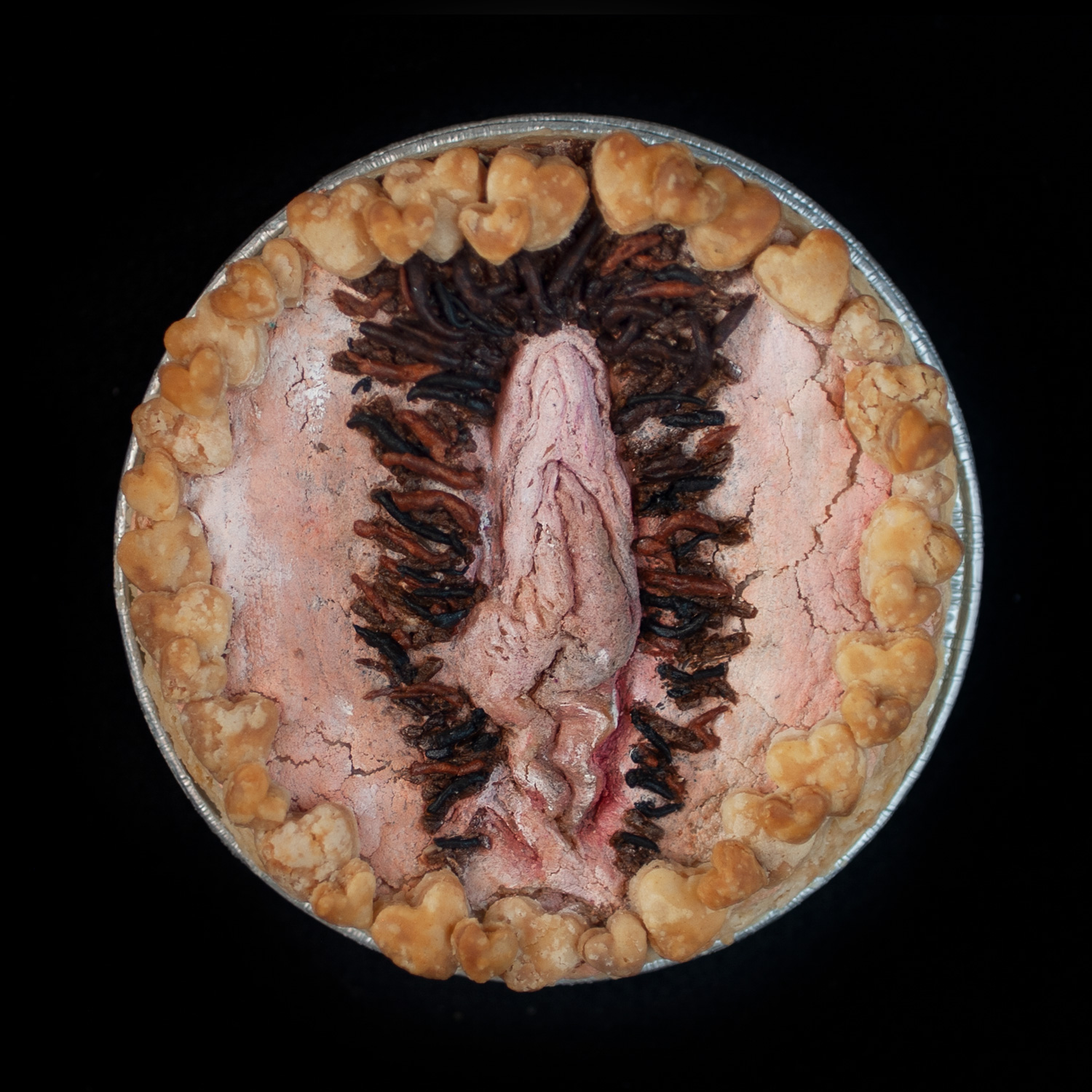 A baked vulva pie on a black background. The pie has brown pubic hair and is surrounded my pie crust hearts. The vulva art is painted to look like a realistic vulva.