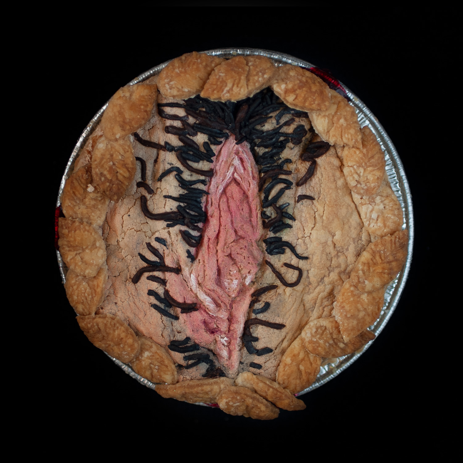 A vulva pie for Pies in the Window on a black background. The Pie features hand-sculpted pie crust art and pubic hair surrounded my pie dough leaves. 