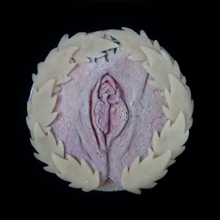 A vulva pie on a black background. The pie is surrounded with pie crust leaves and is painted to look like a realistic vulva.