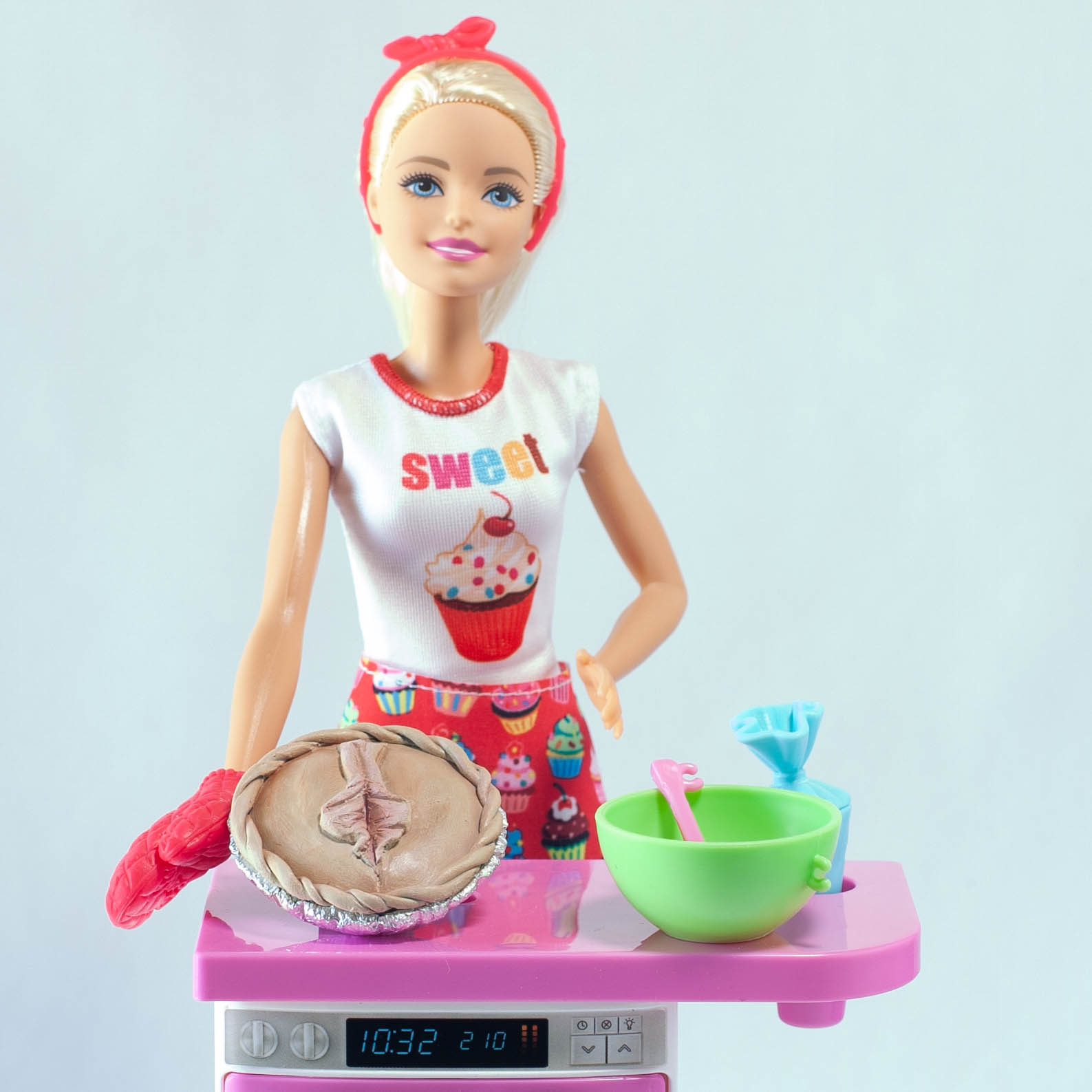 Baking Barbie from the waist up. Barbie is wearing a white t-shirt with a cupcake on it, standing behind a Barbie oven and countertop. On the counter is a vulva pie, mixing bowl, and piping bag. 