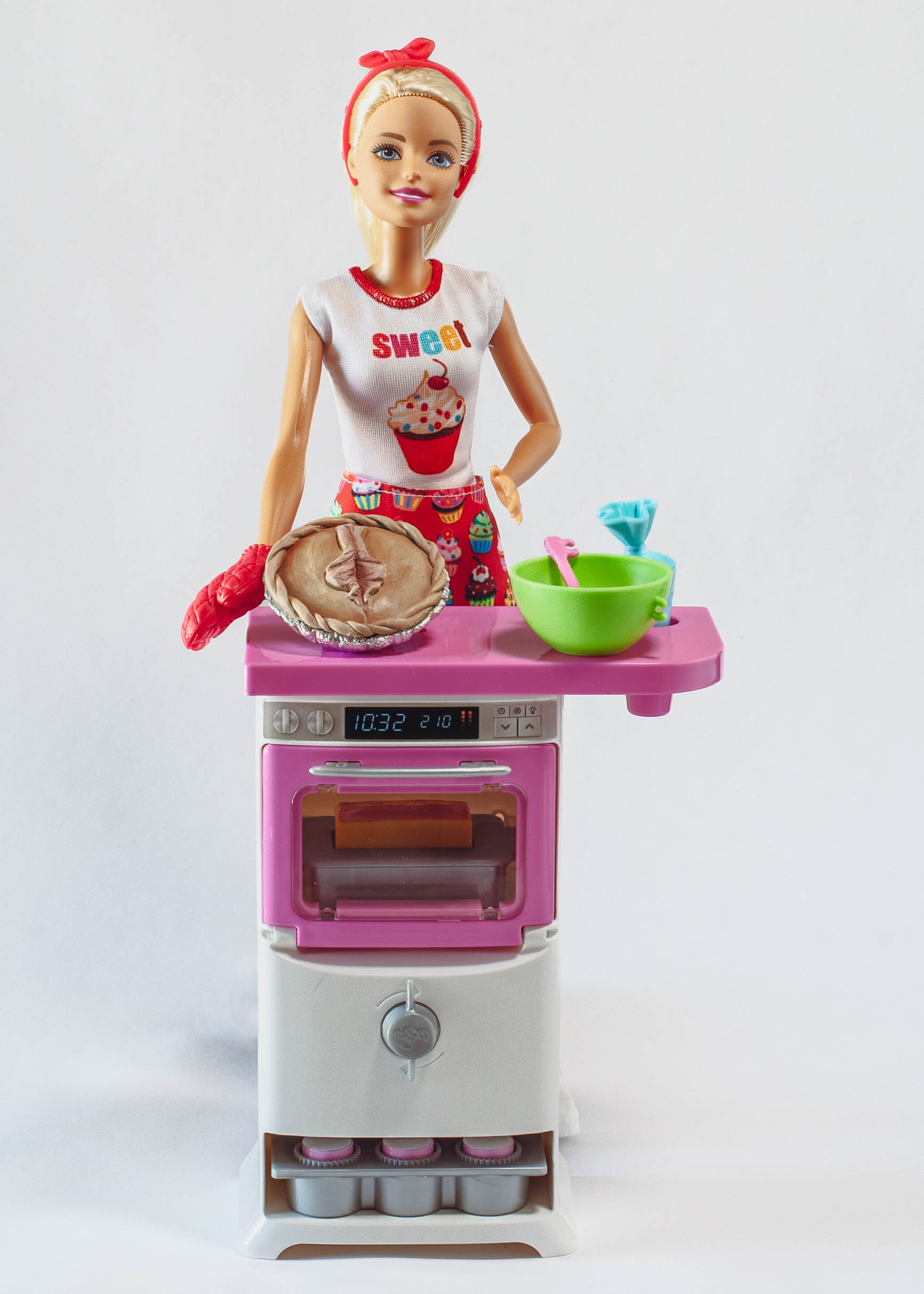 Baking Barbie on a white background. On the counter is a realist vulva pie made from polymer clay and the playset bowl and piping bag. 
