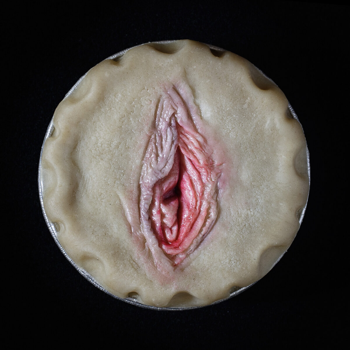 Unbaked vulva pie on a black background. The pie art features a realistic vulva painted with edible paint.