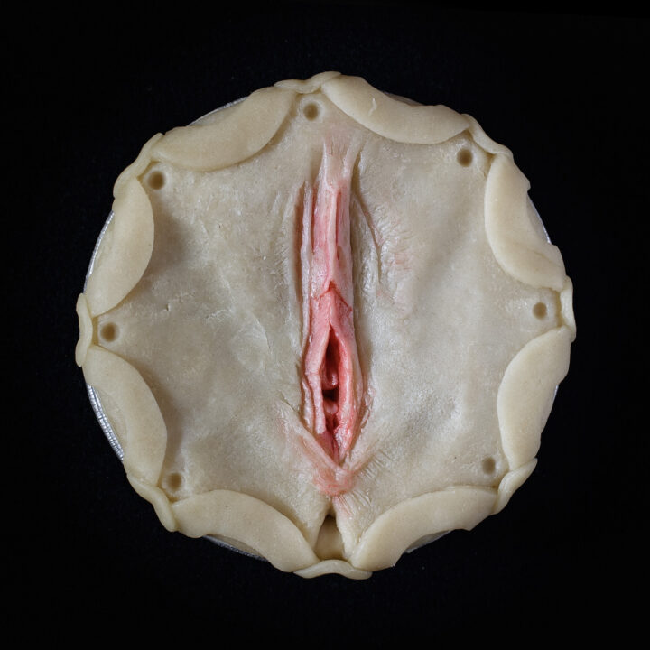 Unbaked vulva pie on a black background, with painted and sculpted realistic vulva