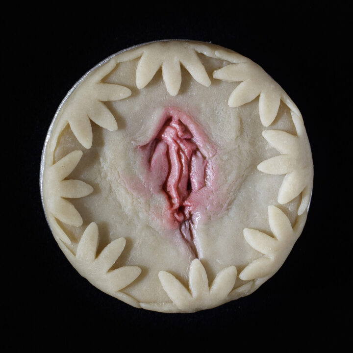 An unbaked vulva pie on a black background. The pie is decorated with cut out flowers and hand sculpted vulva art, painted realistically.