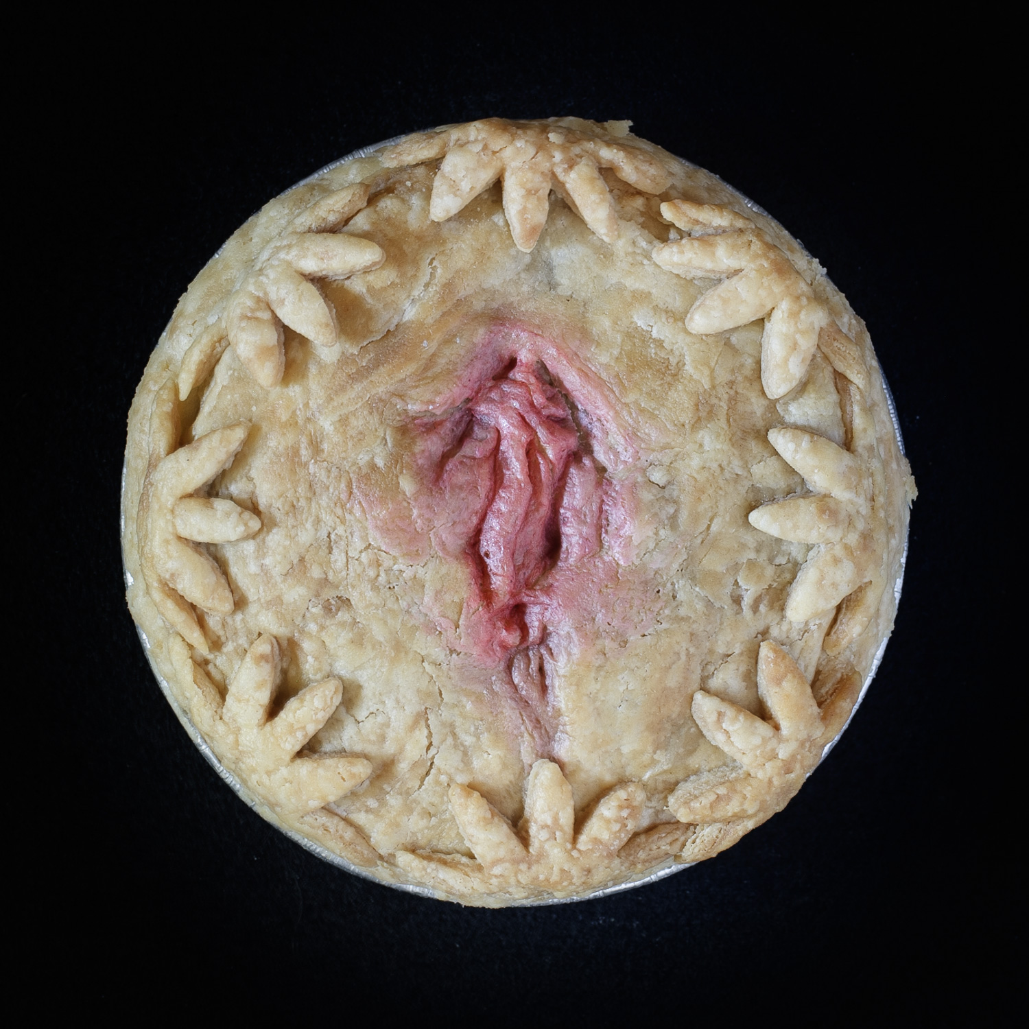 A baked vulva pie on a black background. The pie is decorated with cut out flowers and hand sculpted vulva art, painted realistically.