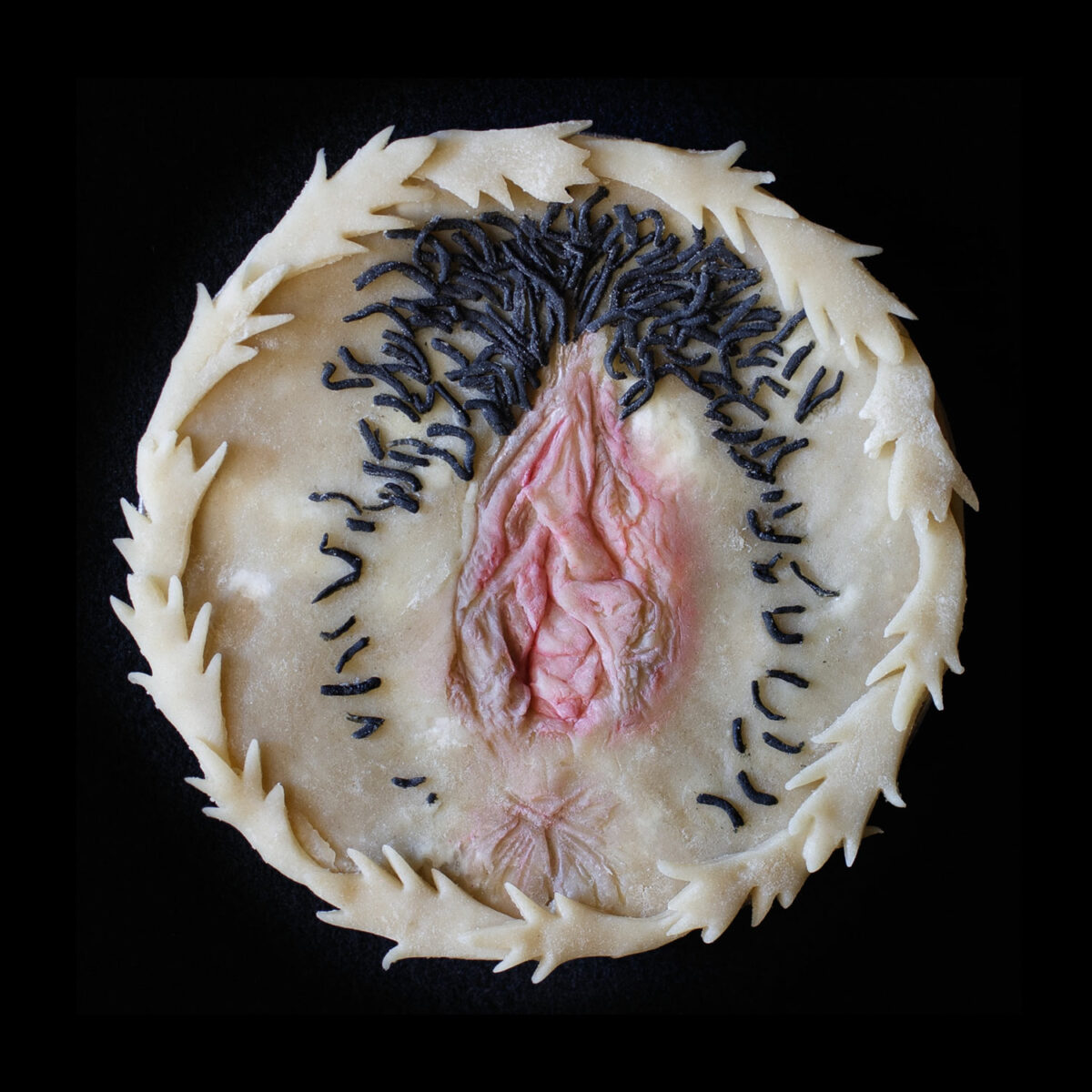 Unbaked version of Pie 73, pie top is decorated with an anatomically correct vulva and hand painted with realistic colors. The pie is on a black background.