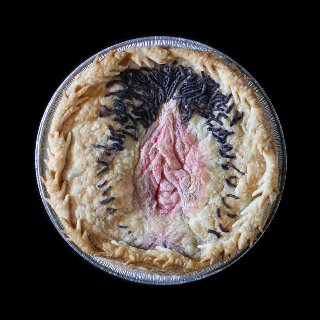 Baked version of Pie 73, pie top is decorated with an anatomically correct vulva and hand painted with realistic colors. The pie is on a black background. 