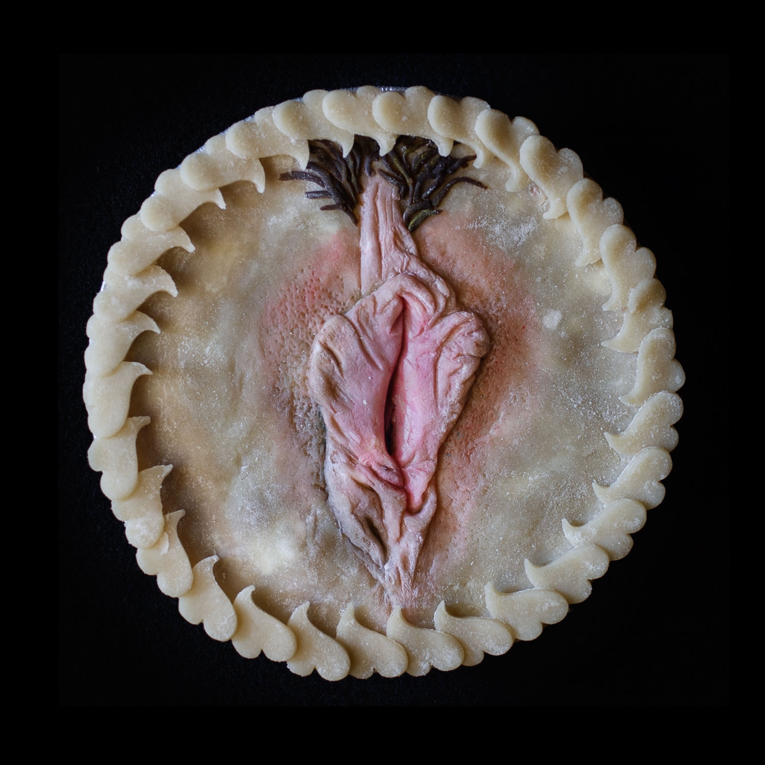 A pie on a black background. The pie is adorned with a realistic vulva surrounded my hearts cut out of pie dough. 