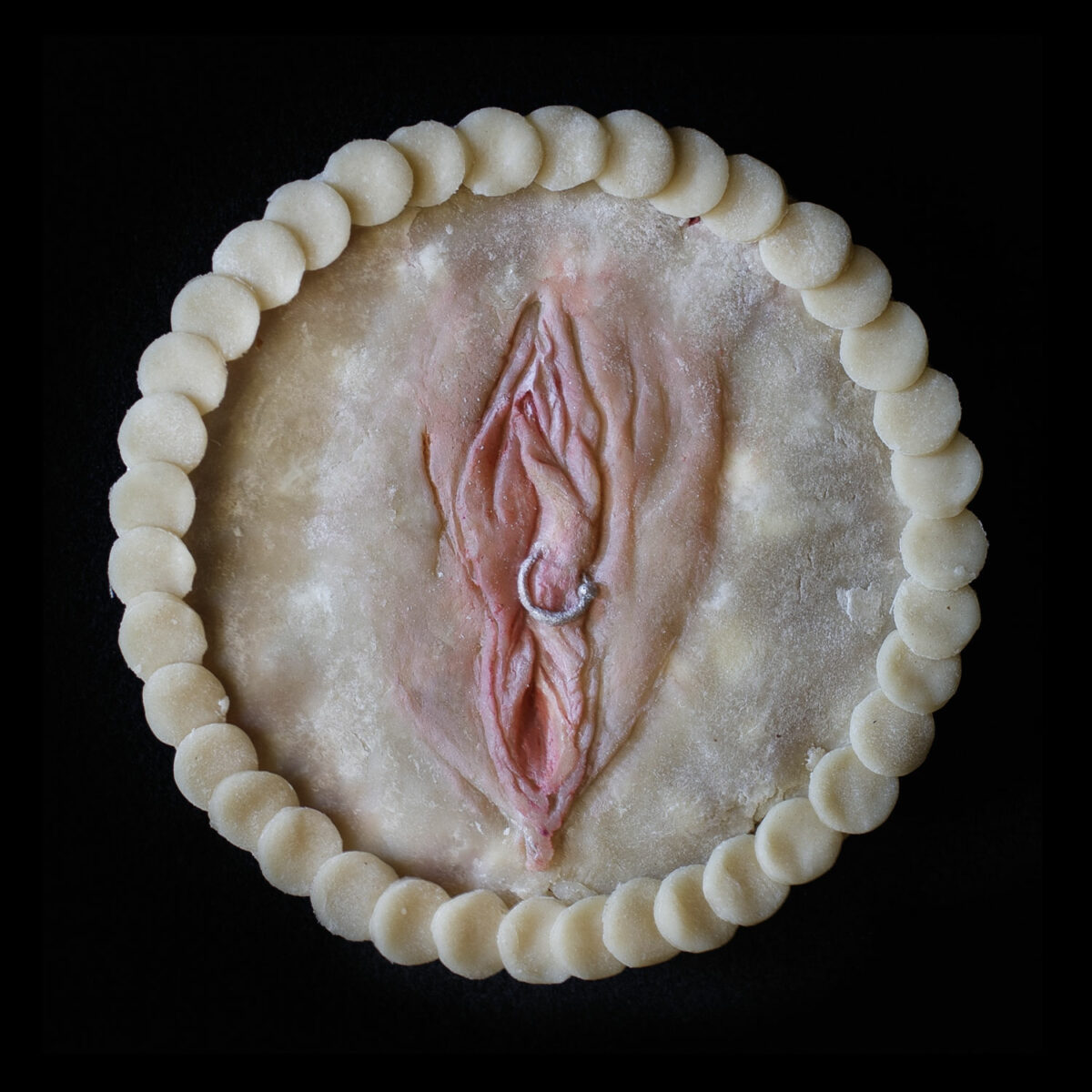 Pie created in the likeness of a vulva. The pie in on a black background with overlapping pie crust circles for the border. The vulva has a silver piercing through the right labia made of pie dough.