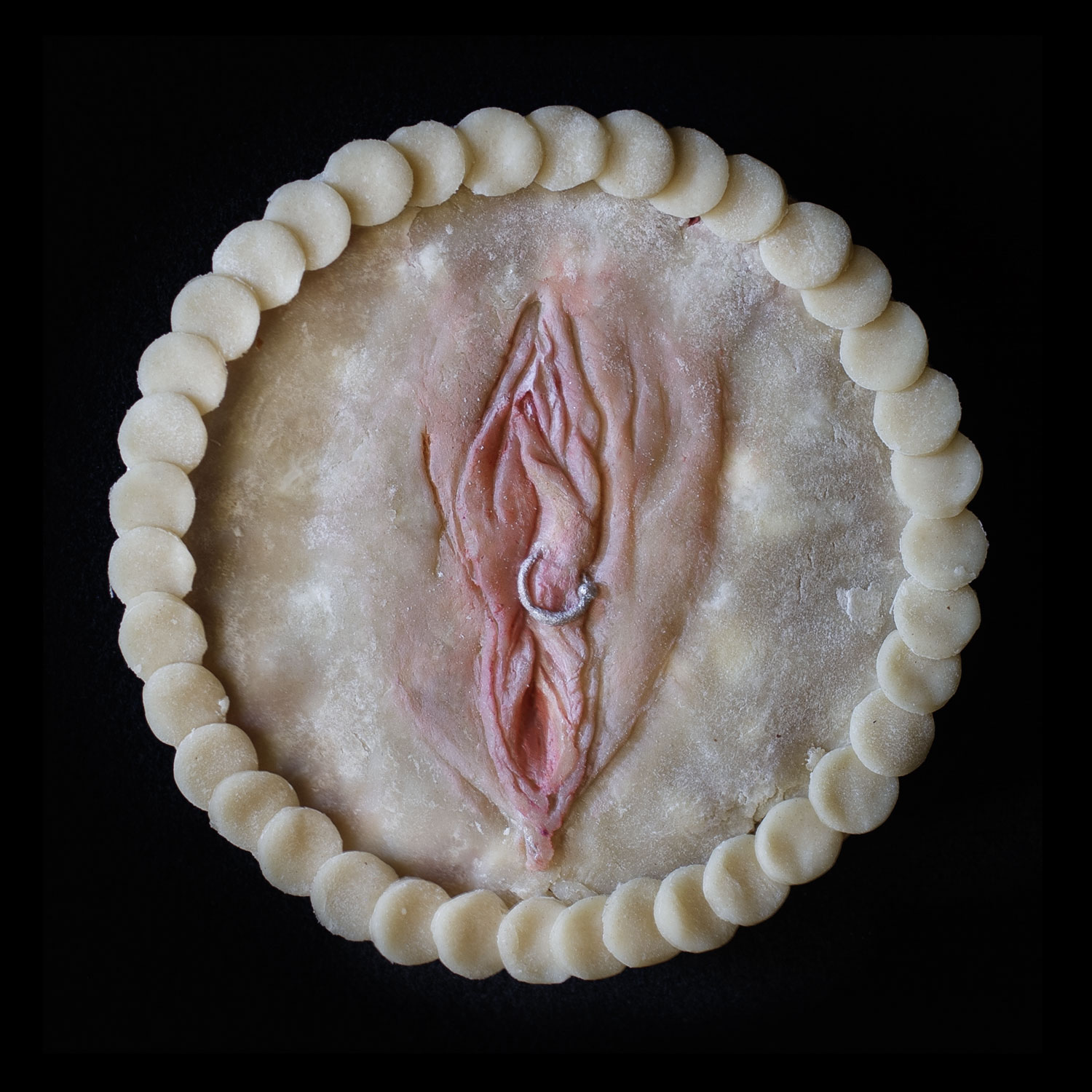 Pie created in the likeness of a vulva. The pie in on a black background with overlapping pie crust circles for the border. The vulva has a silver piercing through the right labia made of pie dough. 