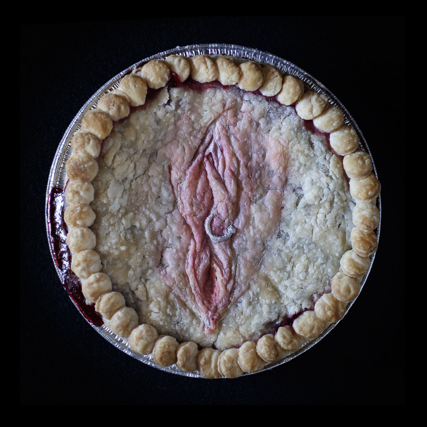 Baked version of pie 75 created in the likeness of a vulva. The pie in on a black background with overlapping pie crust circles for the border. The vulva has a silver piercing through the right labia made of pie dough. 