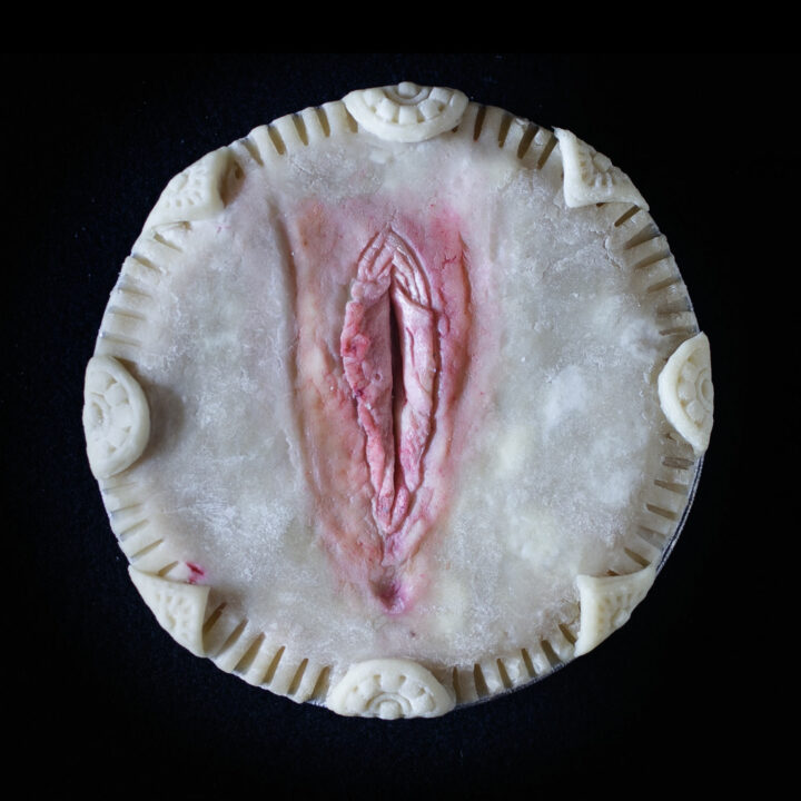 Unbaked pie with hand sculpted art on a black background. The pie art looks like a realistic vulva.
